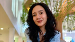 AA26QcX9.jpg Seorang wanita berambut hitam panjang mengenakan kemeja biru muda tersenyum ke arah kamera